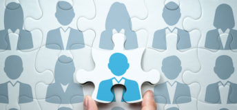A puzzle made up of the grey silhouettes of businesspeople. A hand is placing the final puzzle piece, which has a blue silhouette